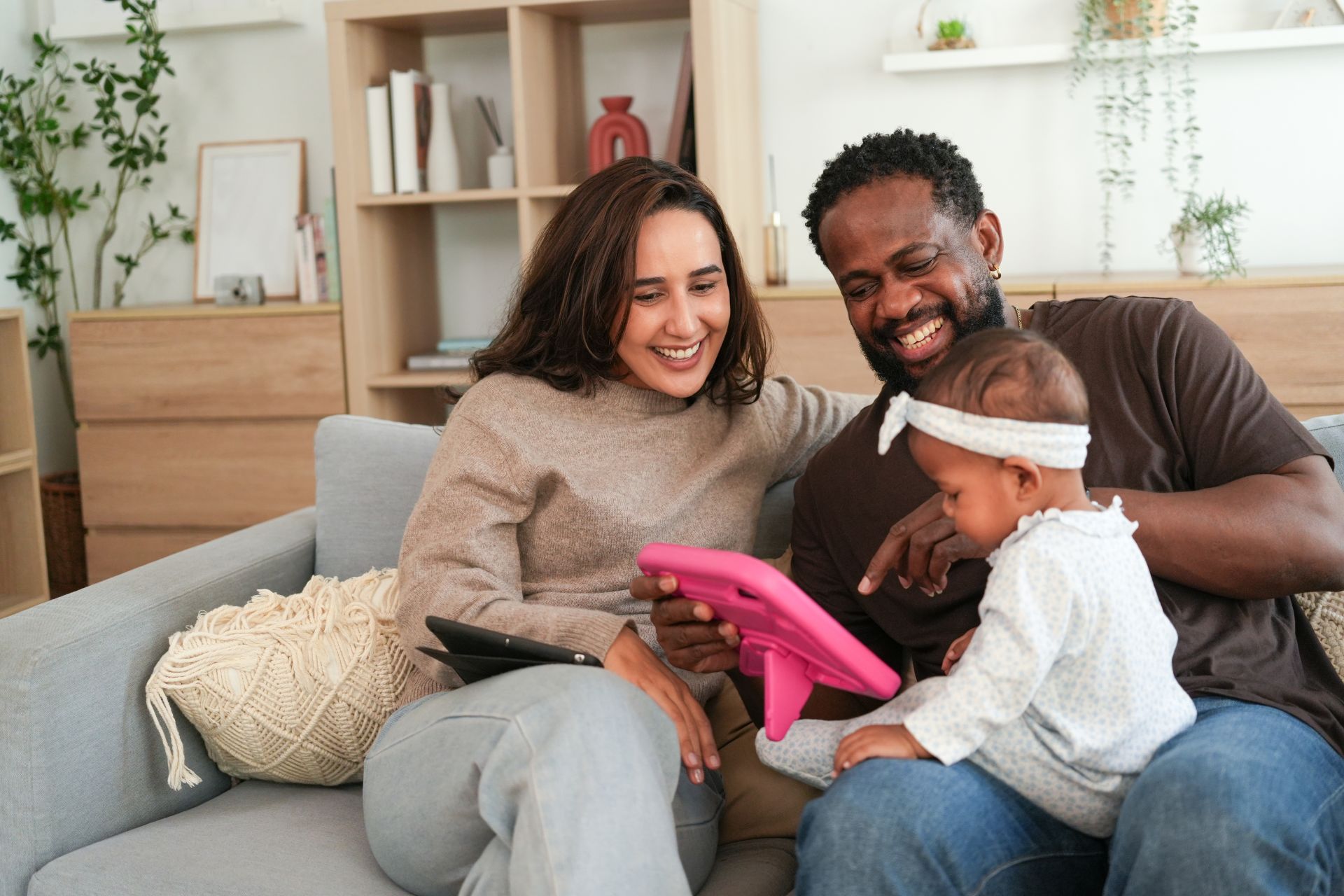 Cheerful multiracial father and mother sitting on a sofa while using a tablet in a child-proof case with their baby daughter. Concept of modern family and home entertainment.