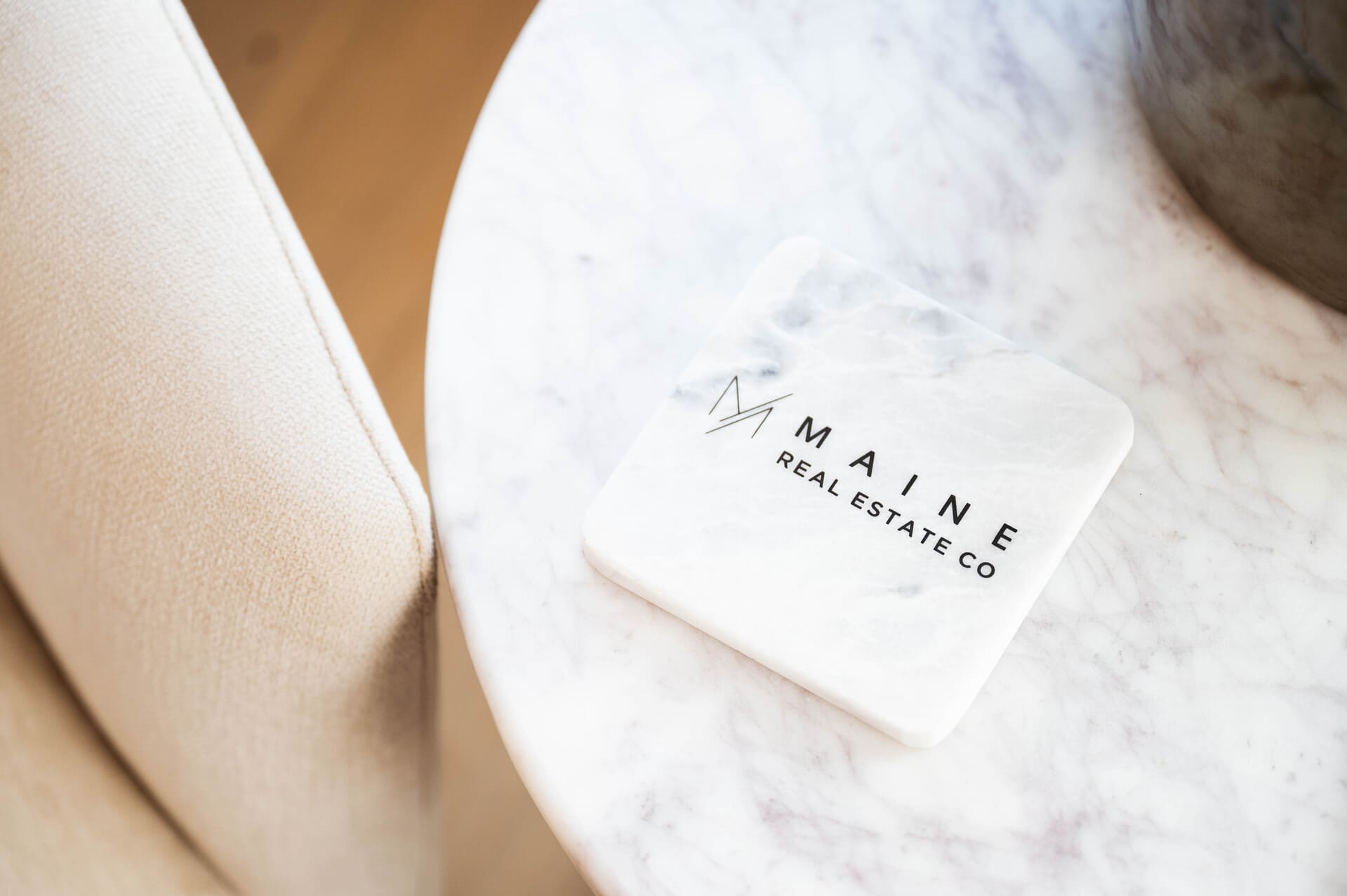 Elegant marble coaster with 'Maine Real Estate Co' logo placed on a stylish white marble table next to a beige textured chair, symbolizing sophistication and modern design in real estate branding.